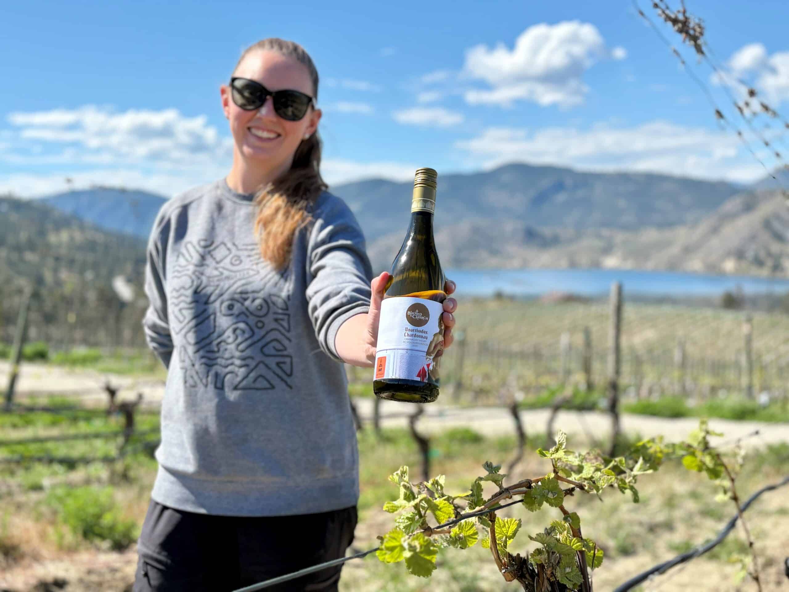 Assistant Winemaker Jamie MacNeill holds a bottle of Unorthodox Chardonnay among the Chardonnay Musque vines at Blasted Church vineyard in OK Falls